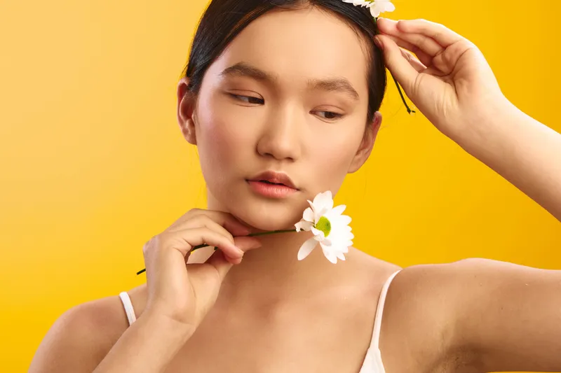 asian woman adjusts a white flower in her hair against a bright yellow studio backdrop, radiating calm elegance and soft beauty with a minimalist, serene portrait.