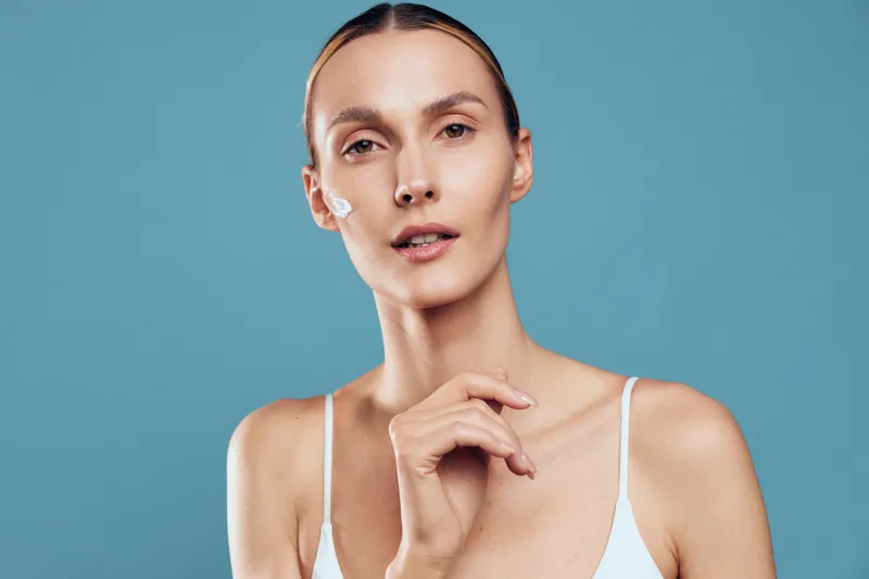 Young woman with clear skin applying white face cream on cheek, wearing white top against blue background. Concept of skincare, beauty routine, and healthy skin care.