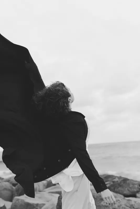 Couple embracing by the seaside, lifting mood and connection as waves crash on rocks, black and white photography capturing romance, motion and shared joy in a coastal moment