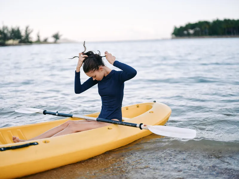 A woman in a navy wetsuit sits in a yellow kayak at the shore, tying her hair as gentle waves roll in, with distant trees framing a peaceful outdoor scene.
