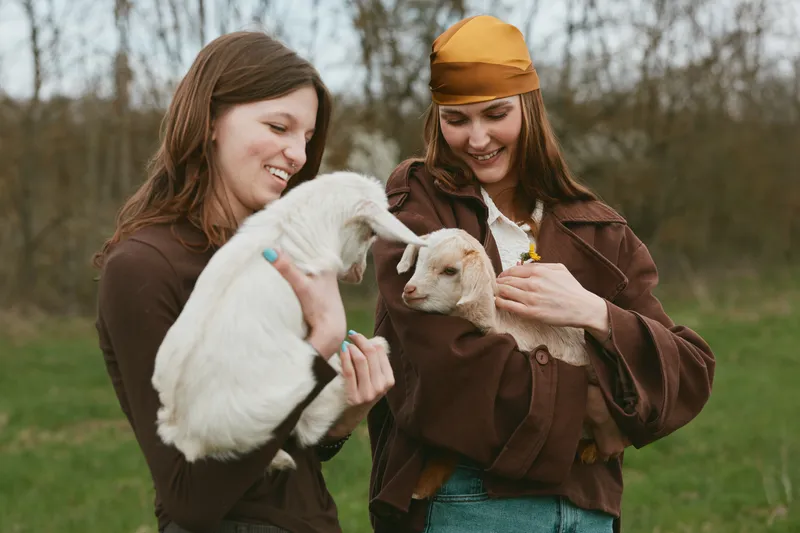 Two women outdoors cradle young goats, sharing a gentle moment of farm life. Casual clothing, natural light, soft grass backdrop. Focus on animal companionship, rural lifestyle, and simple happiness.