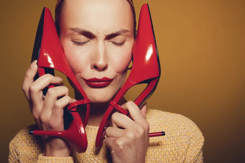 Woman with closed eyes holds red high heels near face, wearing yellow sweater. Concept of fashion, style, emotions, and footwear in studio with warm lighting and brown background.