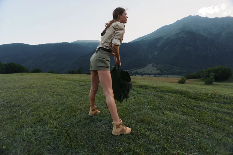 A young woman runs across a field with a mountain backdrop, wearing a light beige top, green shorts and boots, carrying a jacket, conveying freedom and focus.