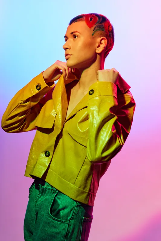 Woman with colorful hair wearing a yellow jacket and green pants posing against vibrant pink and blue gradient background, showcasing modern fashion and bold style.