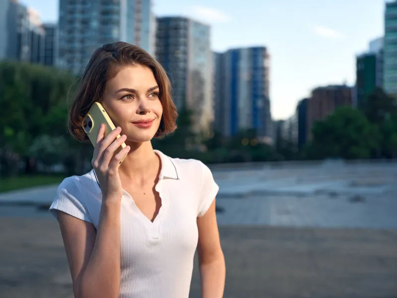 A cheerful professional woman talks on a mobile phone in an outdoor urban setting. She smiles warmly, exuding confidence and focus amid modern buildings and bright daylight.