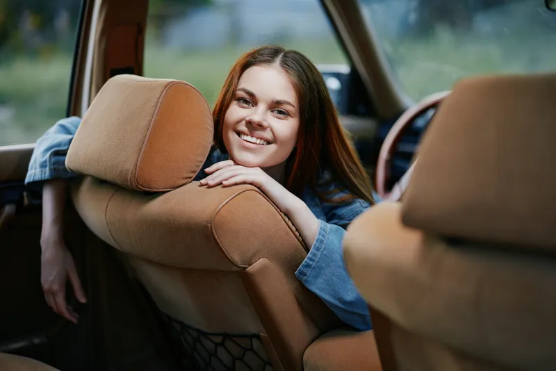 Cheerful young woman in denim jacket smiling from the back seat of a car, enjoying a sunny day outdoors, embodying freedom and adventure, with a warm color palette.