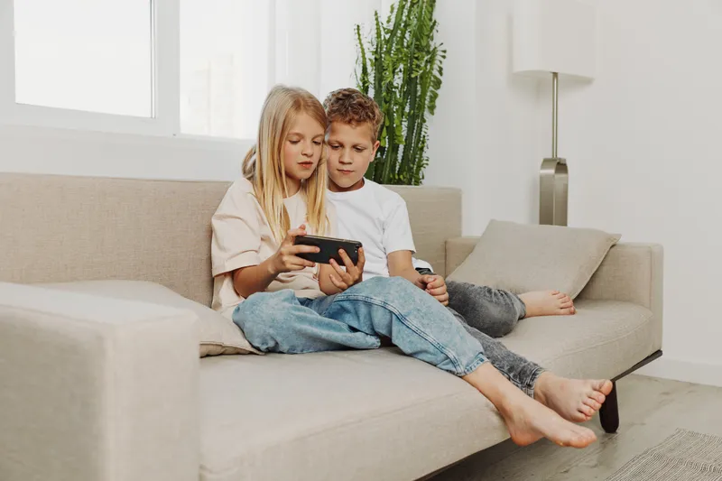 Two kids sitting on a beige sofa watching smartphone together in a bright living room with modern decor and casual clothes.