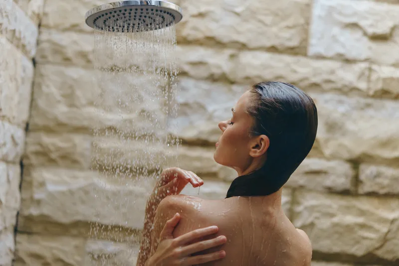 woman showering under rainfall showerhead with stone wall background, wet hair, relaxed, natural skincare, spa atmosphere, wellness concept