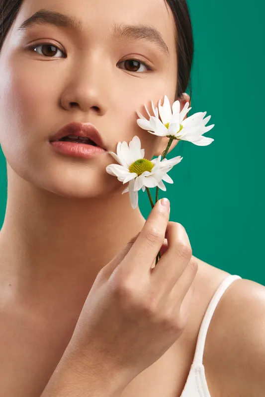 Asian woman portrait holding white daisies against a vibrant green background, exuding calm beauty and subtle emotion, creating a fresh, natural mood with soft light and gentle detail.