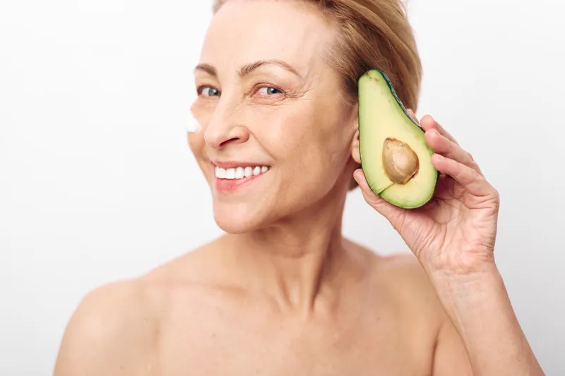 Smiling mature woman holds half avocado near face with healthy skin and natural beauty on white background. Concept of wellness, nutrition, and organic skincare for aging skin.