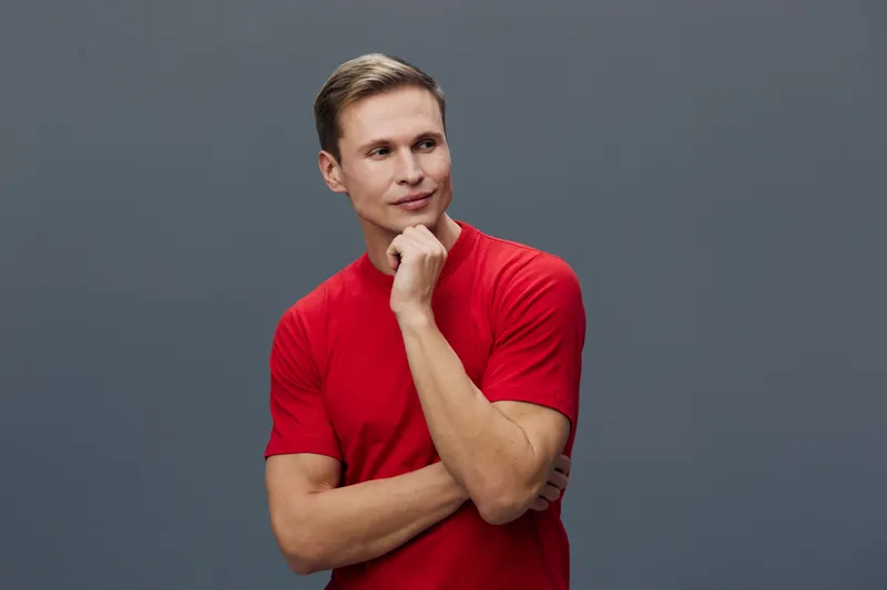 Thoughtful young man with short brown hair, wearing casual red t-shirt, standing against a plain grey background, posing with hand on chin in a studio portrait. People lifestyle concept.