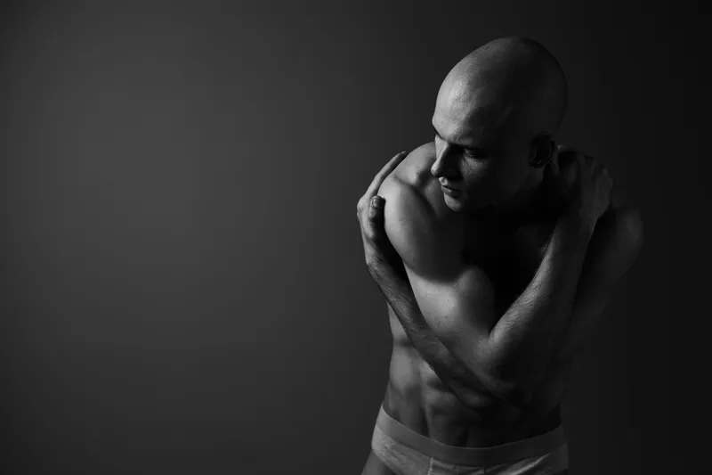 A contemplative man poses gracefully in a black and white setting, showcasing strong muscles and a reflective mood with an artistic touch.