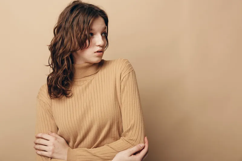 Young woman with curly hair wearing beige sweater looks sideways with thoughtful expression on plain beige background. Concept of contemplation, casual style, and natural beauty.