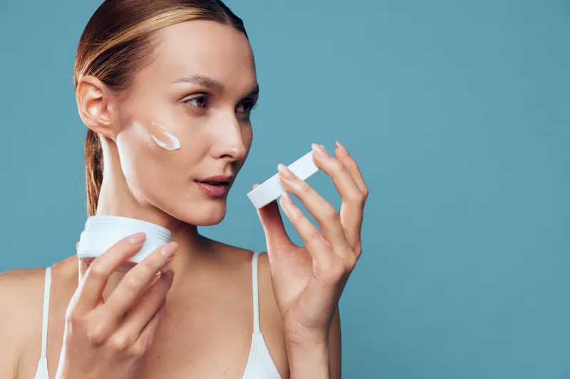 Smiling woman applies white moisturizer on her cheek in a close-up studio shot, blue background, showcasing skincare routine, self care, and healthy glow with soft lighting.