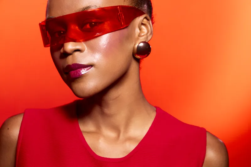 Stylish African American woman with dark skin wearing red sunglasses and red top poses against orange background. Concept of fashion, beauty, and modern accessories with confident mood.