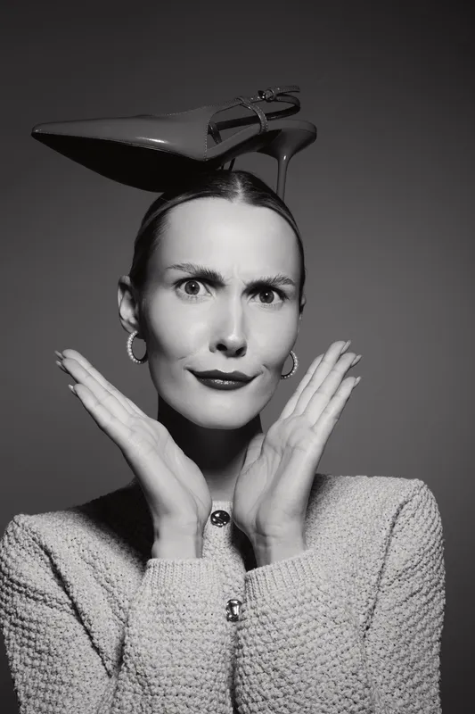 Caucasian woman with light skin wears shoe on head and makes confused face with hands near cheeks in black and white portrait.