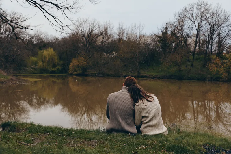 A young couple sits closely together by a calm river in autumn, wearing cozy knitted sweaters and enjoying a quiet moment in nature with bare trees around them.
