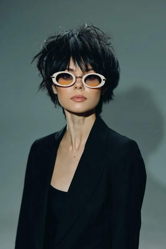 Stylish woman with short hair wearing white sunglasses and black outfit, posing confidently against plain background.