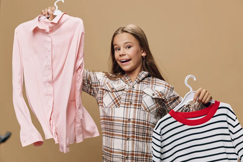 Girl with long hair holding two clothes hangers with shirt and sweater, smiling and wearing plaid shirt on brown background. Fashion choice, style and clothing options for kids.