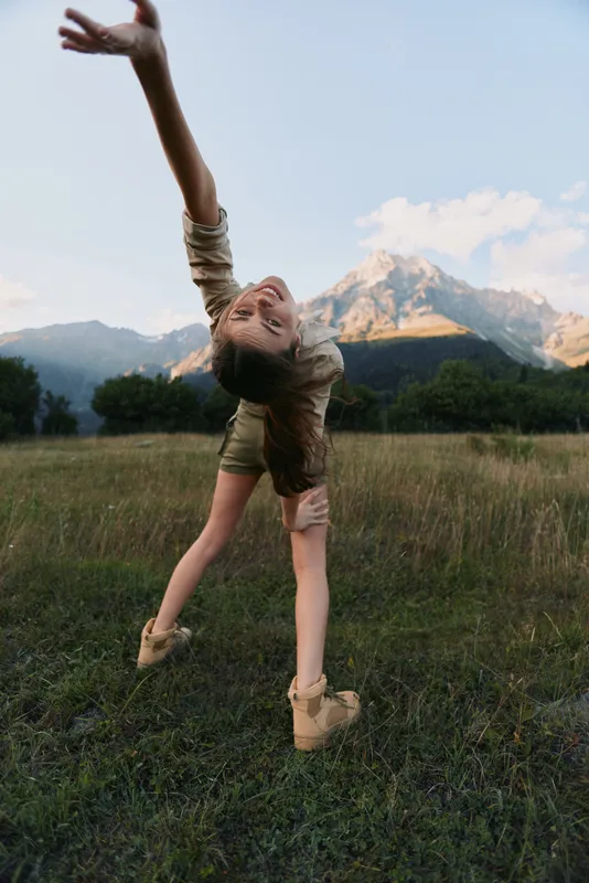 Young woman performs a backbend in a sunlit meadow with distant mountains, capturing a moment of playful energy and balance in a natural outdoor setting as a gentle breeze lifts hair