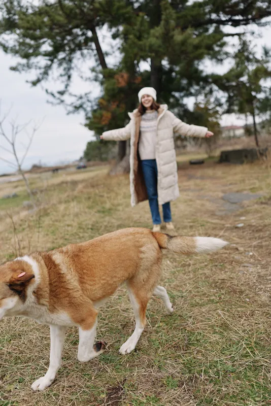 Woman in a light beige coat and knitted hat stands with arms open as a large dog runs through a park on a chilly winter day.