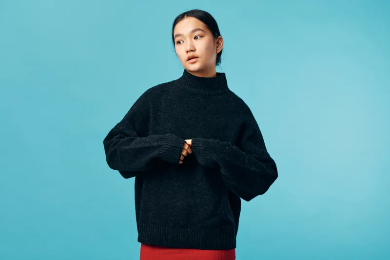 Young woman wearing black oversized sweater standing against blue background looking aside with thoughtful expression and natural makeup for casual style and fashion.