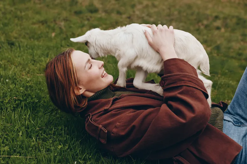A young woman with red hair lies on grass smiling as she holds a white baby goat close to her face in a casual outdoor lifestyle moment.