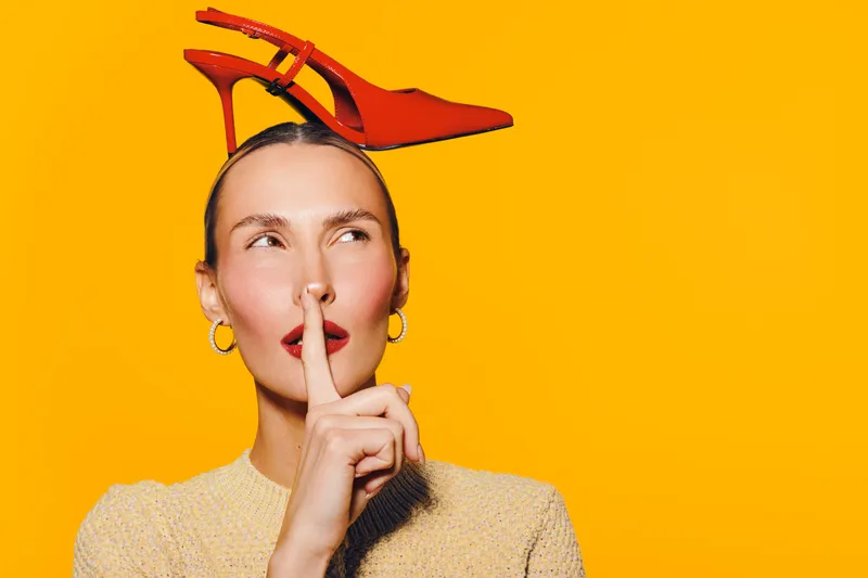 Young woman with red high heel shoe on head holds finger to lips in shushing gesture. Bright yellow background creates bold contrast. Concept of fashion, silence, and playful expression.