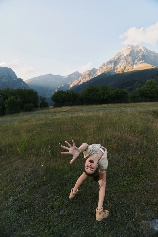 Energetic woman bends toward the camera in a sunlit meadow, mountains in the background, casual outdoor wear, capturing a playful and adventurous lifestyle moment