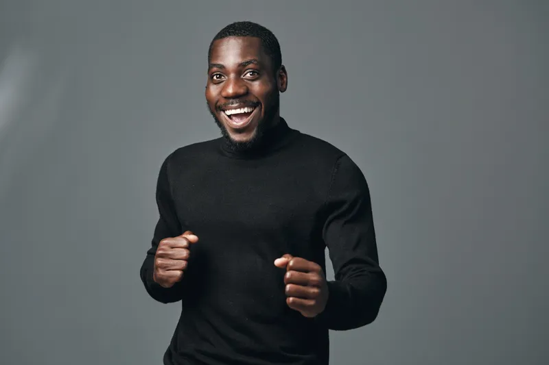 African man smiling and posing with fists up in creative conceptual visual style studio portrait. Happy black male model with positive energy and confident expression on gray background.