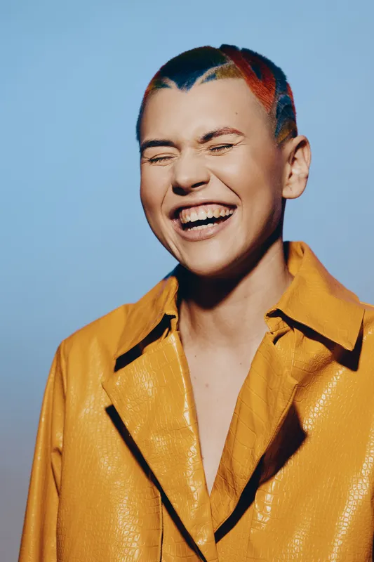 Laughing woman with colorful short hair wearing a bright yellow textured jacket against a soft blue background, expressing joy and individuality in fashion.