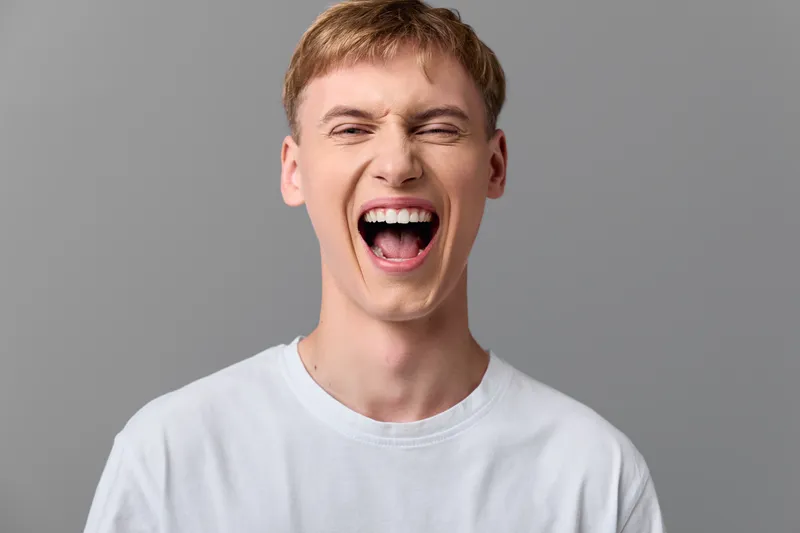 Laughter and open mouth expression by a young man in a white tee, conveying energy, excitement, and cheerful emotion against a neutral studio background.