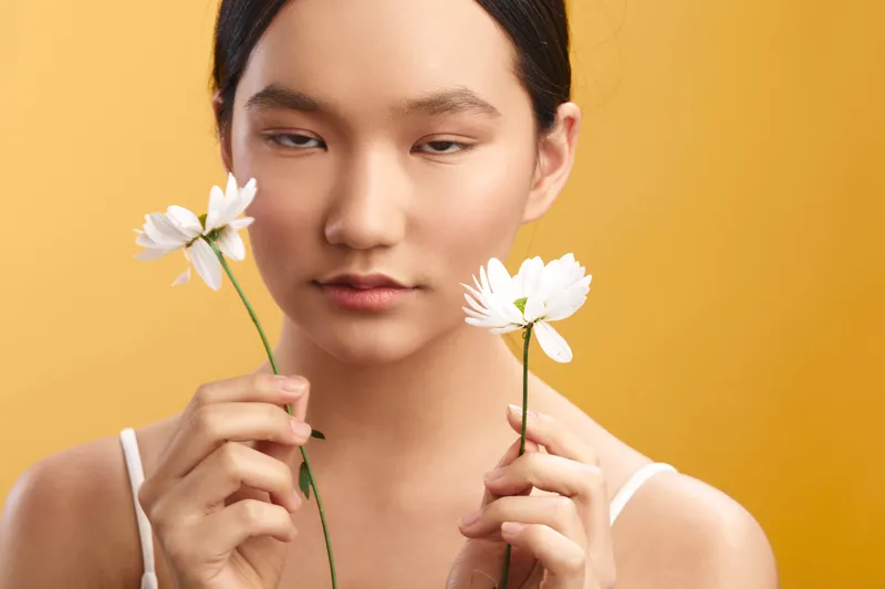 Asian woman gently holds white flowers against a warm yellow backdrop, her calm expression inviting viewers to sense serenity, beauty, and quiet confidence.