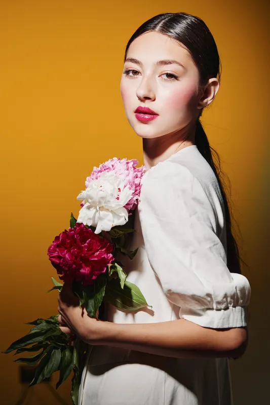 Young female beauty model with long sleek hair holds vibrant pink and white peonies against orange background. Soft makeup and elegant blouse create fresh fashion concept.
