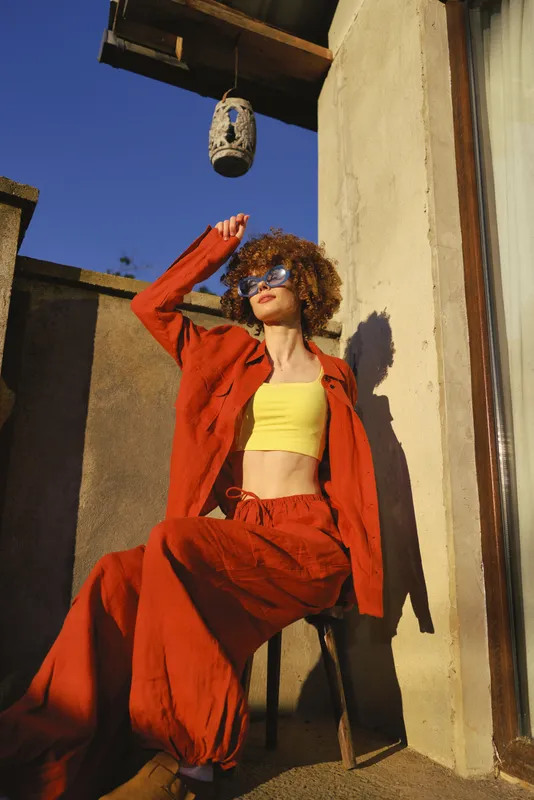 Confident woman with curly hair wearing a yellow crop top and red outfit lounges on a sunny terrace, showcasing bold fashion and relaxed lifestyle in warm light