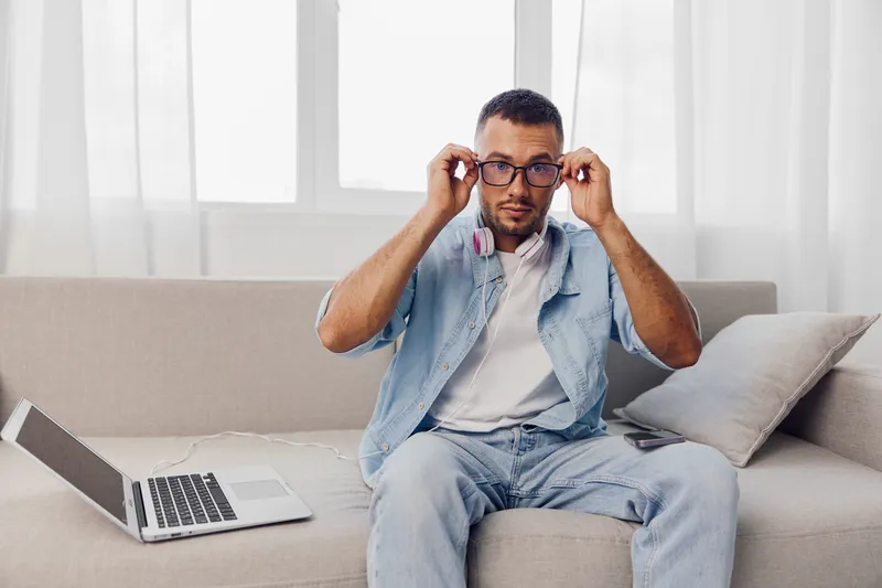 Focused man adjusting his glasses while sitting on a stylish sofa, surrounded by natural light. This trendy scene highlights modern lifestyle and remote work.