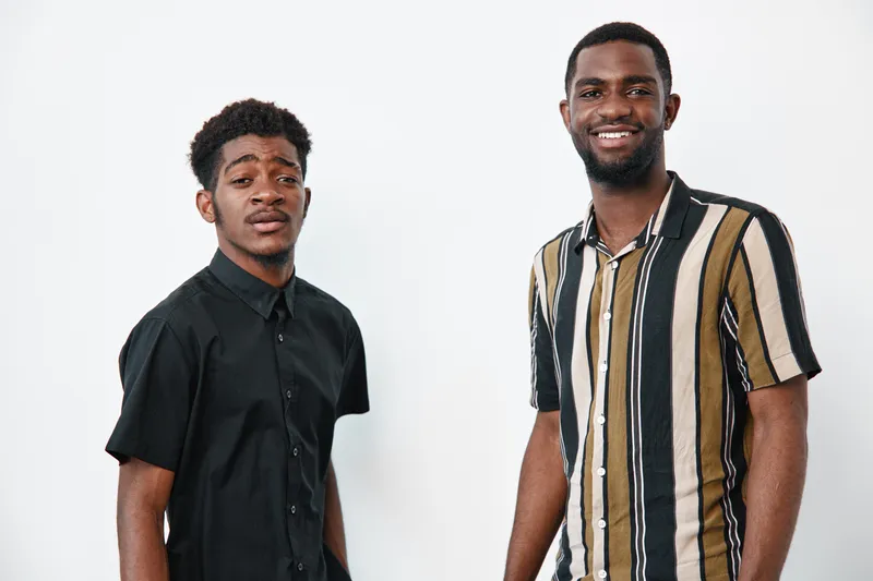 Two young African American men with dark skin standing and smiling in casual shirts against white background. Portrait of confident friends with diverse style and positive mood.