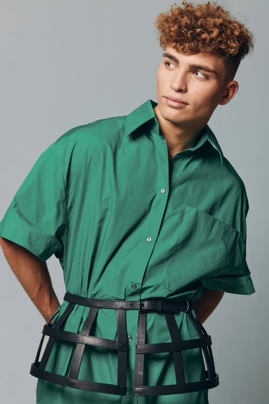 Young man with curly hair wearing green shirt and black leather cage belt posing against gray background in studio for fashion portrait and style concept.