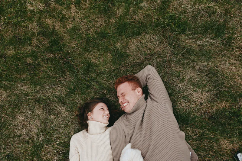 Couple lying on grass outdoors, enjoying a relaxed moment together, smiling and cuddling under soft sunlight, cozy sweaters, natural colors, lush greenery background, peaceful lifestyle scene