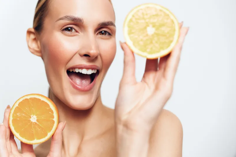 Happy woman holding two halves of fresh orange close to her face, smiling with open mouth, clear skin, bright background, healthy lifestyle concept, natural fruit