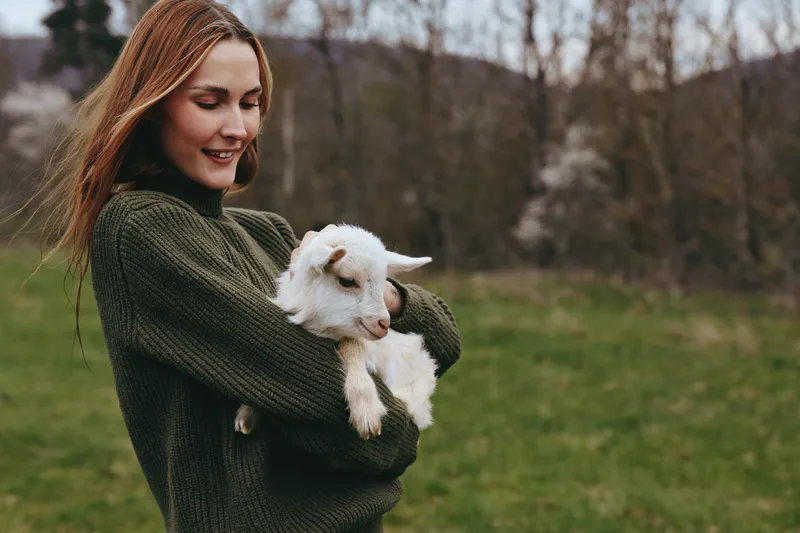 A woman in a dark green sweater holds a fluffy white goat kid in a green field, presenting a warm countryside lifestyle moment with gentle touch and peaceful animal companionship