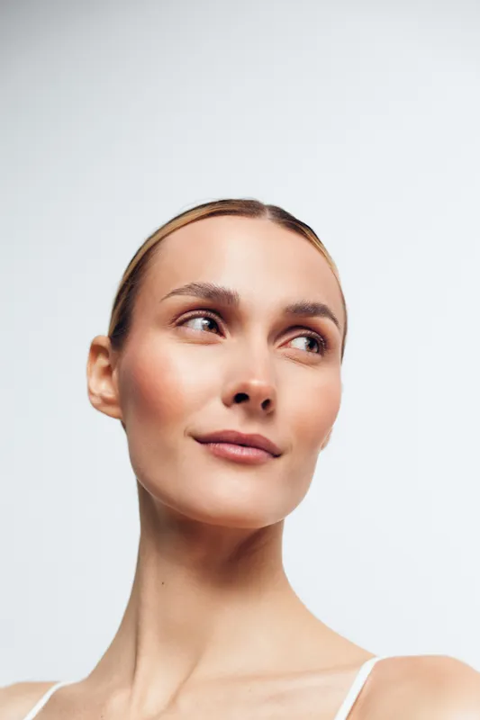 Young woman with blonde hair gazes upward in a clean studio portrait, soft lighting highlighting her calm, confident expression and smooth skin against a neutral background.