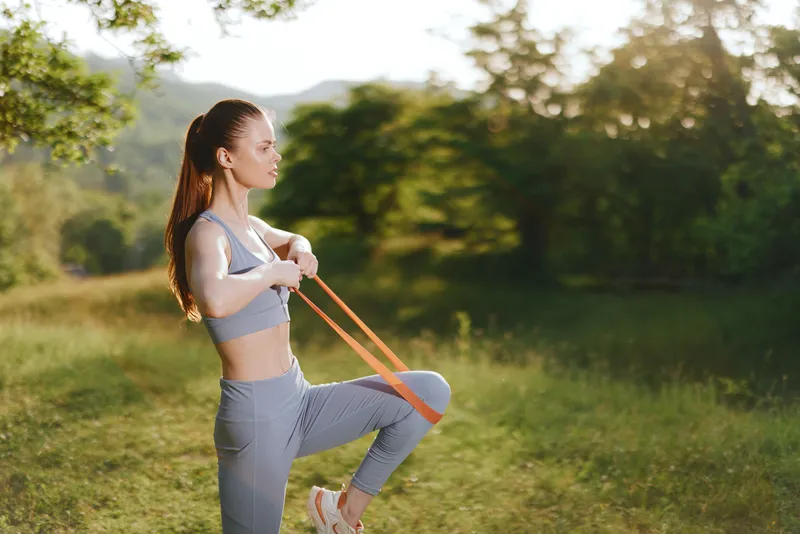 Fit young woman exercising outdoors with resistance bands in a sunny park, showcasing healthy lifestyle and fitness concept