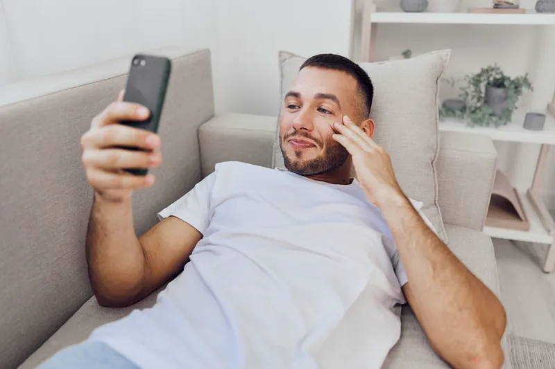 Relaxed man using smartphone while lounging on a cozy sofa, enjoying leisure time at home. His expression shows contentment and engagement with the device.