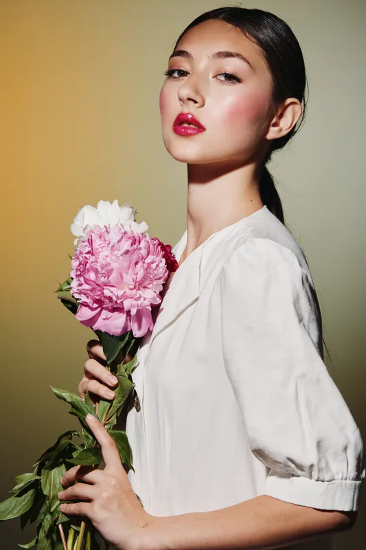 Young woman with glossy dark hair poses with a large pink flower against a gradient background, showcasing natural beauty and modern fashion with vibrant makeup and sleek styling.