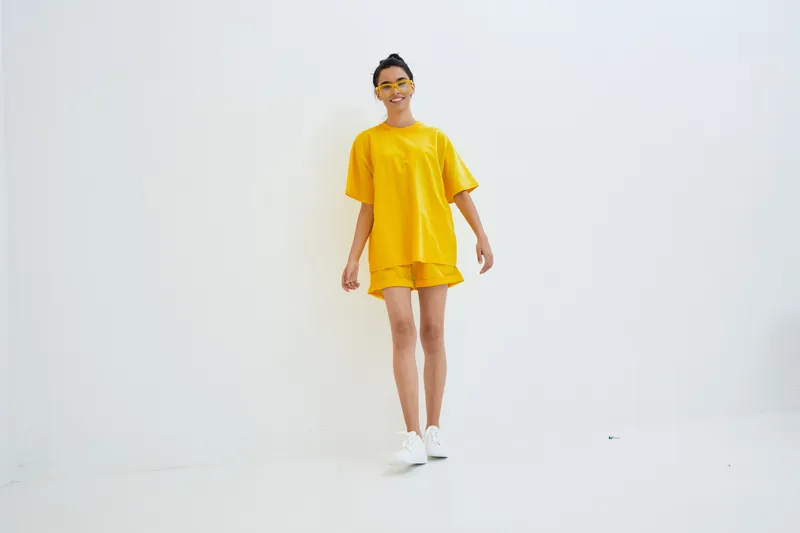 smiling woman wearing bright yellow casual outfit stands in minimalistic white space with sporty sunglasses, radiating confidence and summer vibes in studio lighting