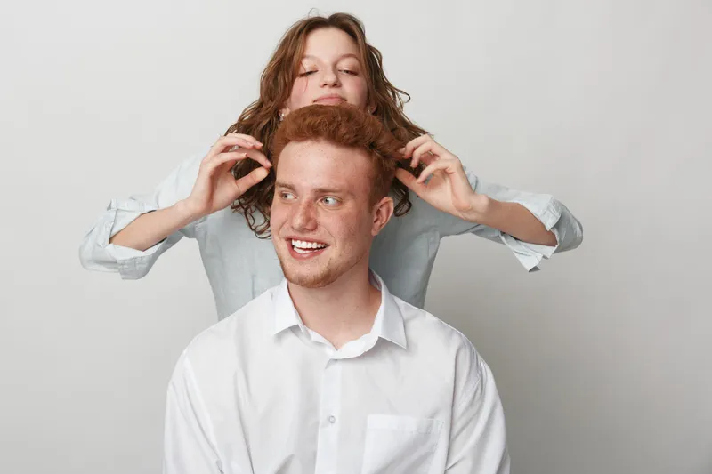 Happy young man with red hair smiles while woman behind him playfully touches his ears. Casual studio portrait on white background with friendly and joyful mood.