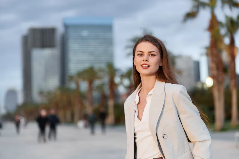 confident woman walking outdoors in business attire near urban skyscrapers with palm trees and cloudy sky during daytime