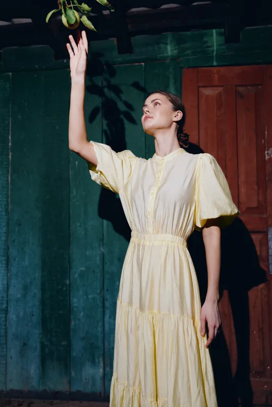 Woman in yellow vintage dress reaching for hanging fruit indoors authenticity inclusivity portrait with dramatic shadow and rustic wall
