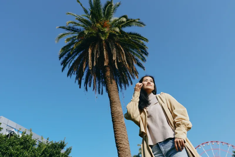 Fashion, outdoor, palm, scenery, casual, model posing beside a tall palm under a clear blue sky, wearing a beige coat and light sweater, urban vibe on a sunny day.
